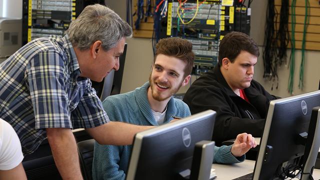 2 smiling students working on a computer as an instructor leans down to help