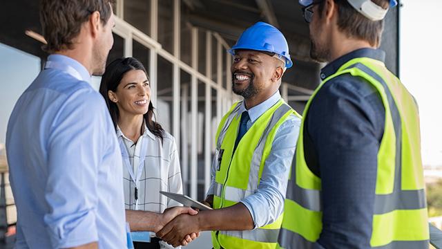 A man in a blue hard hat and safety vest shakes hands with a man in a button down shirt while a woman looks on and smiles.
