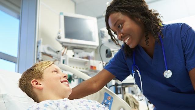 A nurse in blue scrubs smiles at a young boy in a hospital bed.