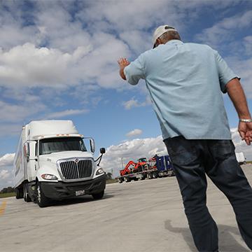 A cdl instructor stands with his back to the camera and hand raised while directing a student driving a semi truck