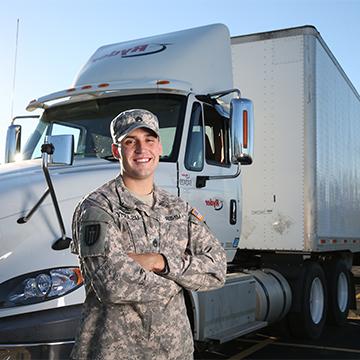 A male veteran in uniform with arms folded looks at the camera and smiles while standing in front of a semi truck