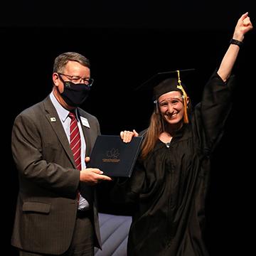 A GED graduate wearing a cap, gown and face shield receives her diploma from 赌钱app可以微信提现 president Dr. Andy Bowne while smiling at the camera and raising a fist in the air