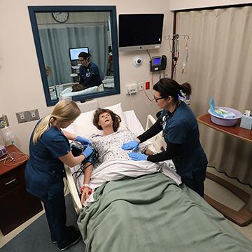 2 nursing students practice care on a simulation dummy while being observed by a nursing faculty member