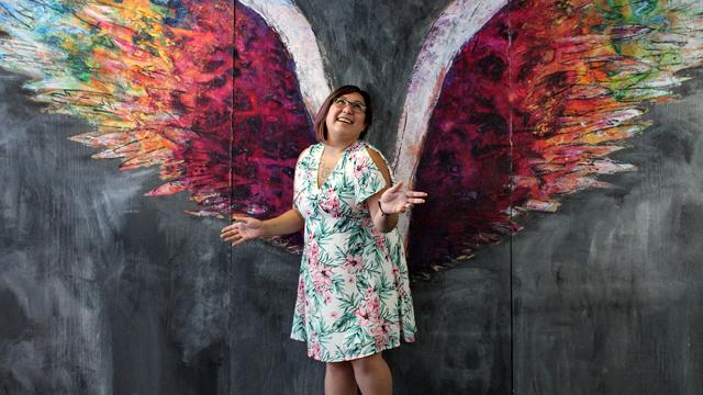 Vanessa McCarron poses in front of a colorful wall mural of angelic colorful wings.