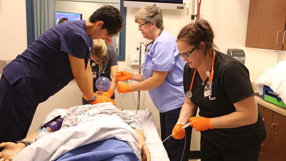 Three students practices aspects of healthcare on a simulation mannequin while an instructor looks on.